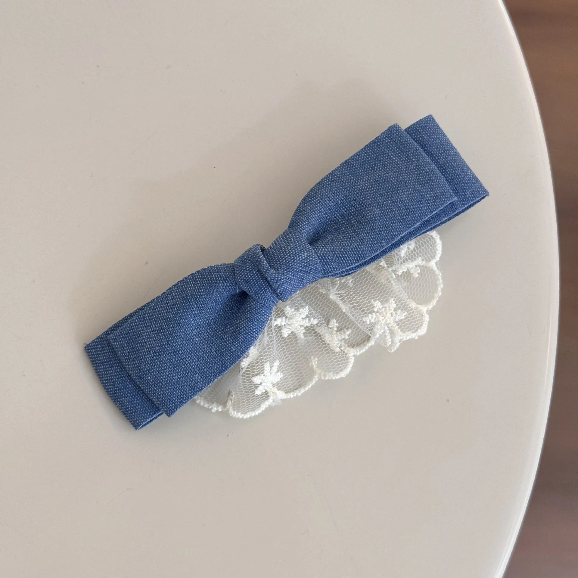 A women's hair clip barrette with a soft blue cloth bow and lace decoration on a plastic clip.