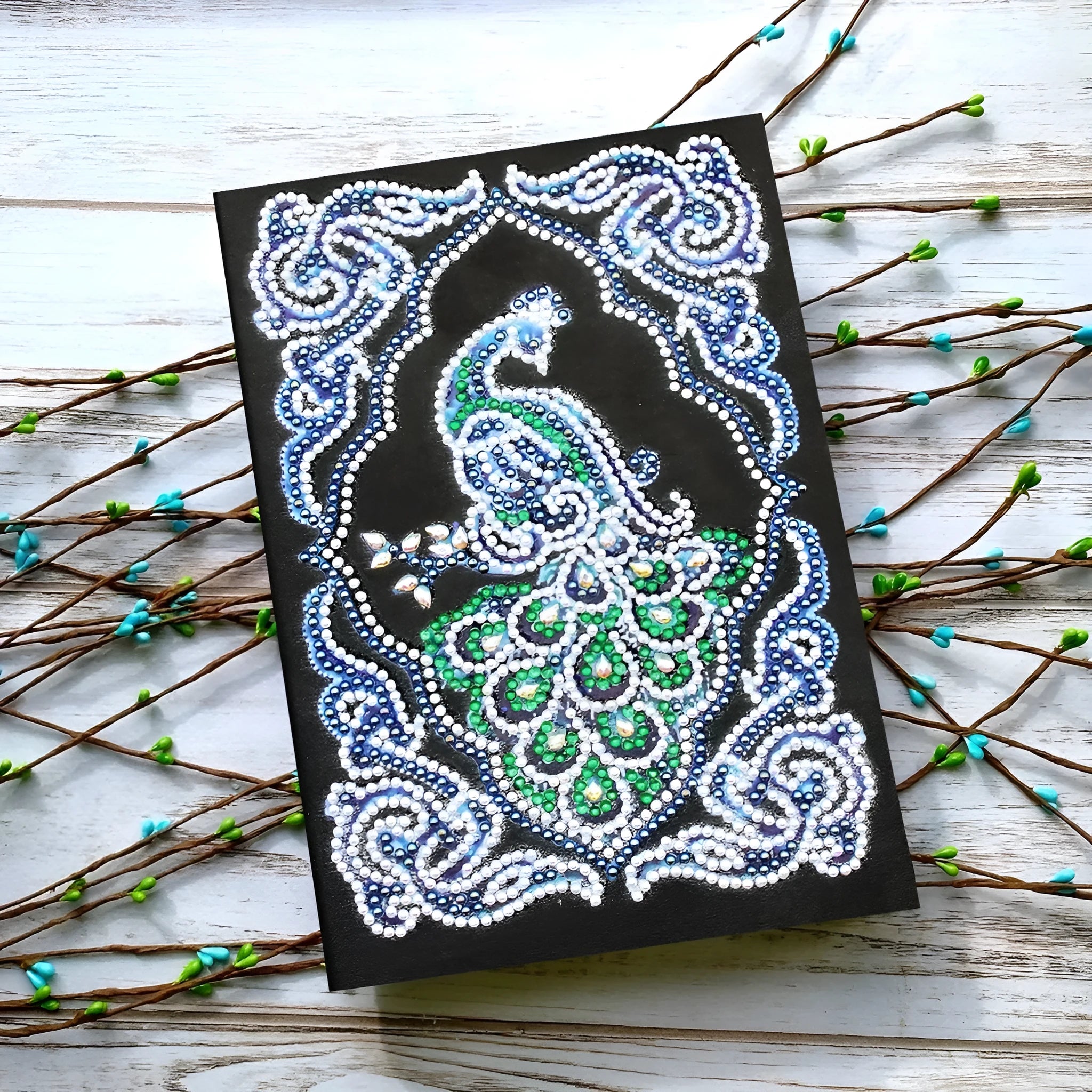 Black A5 diamond painting notebook with peacock design, open to show shaped and bright drills on page.