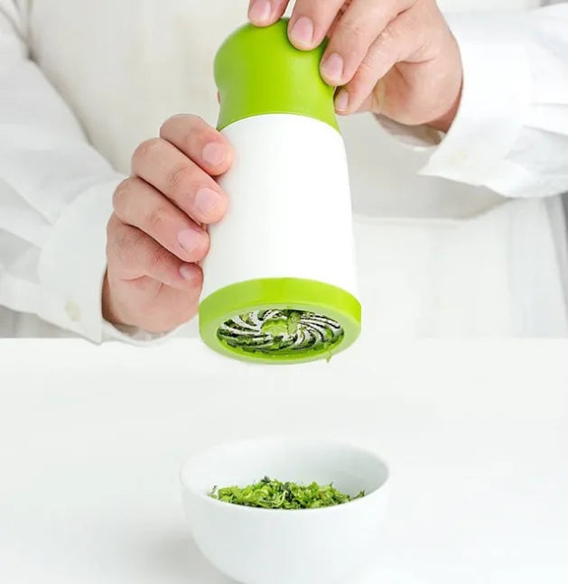 White and grey manual herb grinder with stainless steel blades and removable top on a kitchen counter.