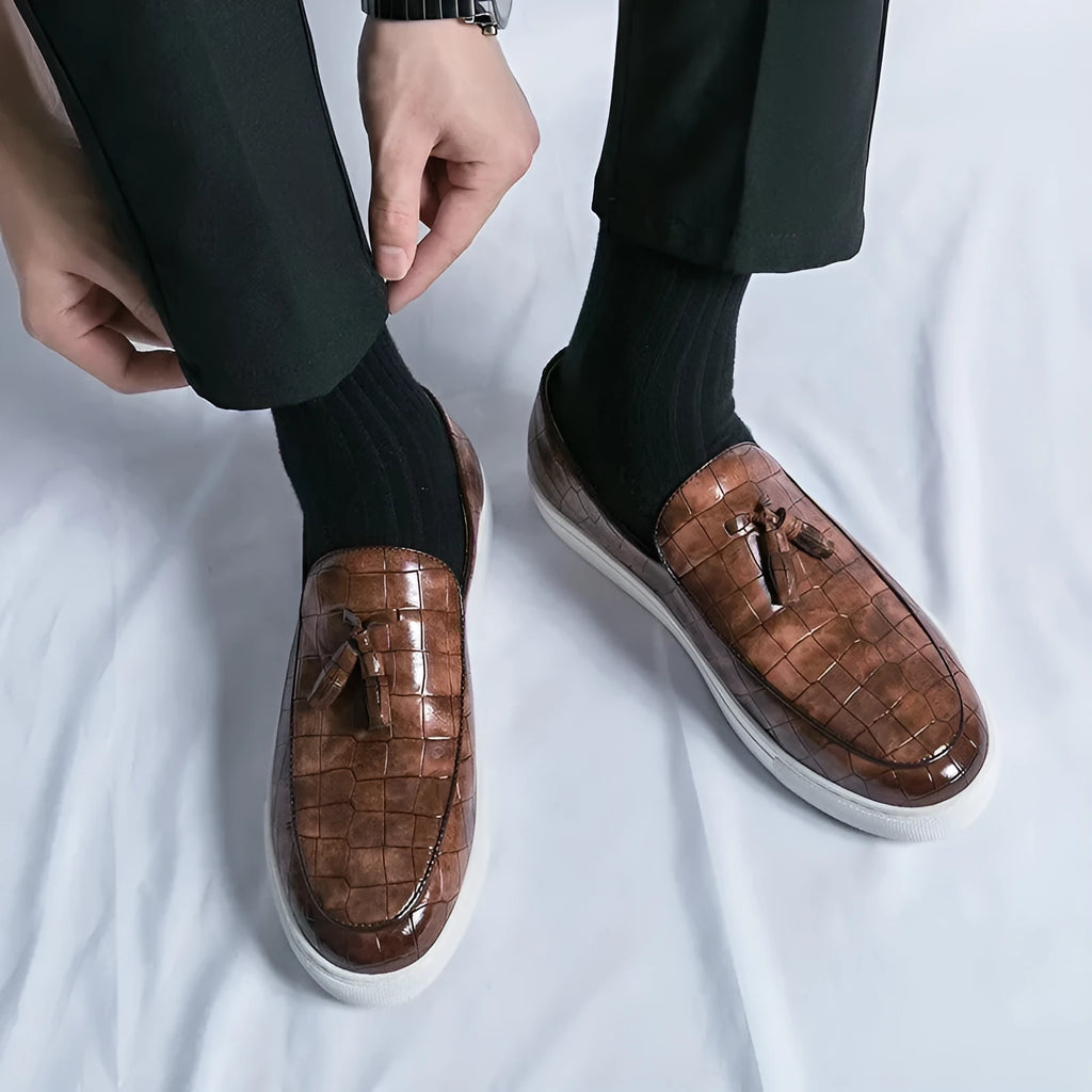 Men's black smart-casual sneakers with a tassel detail on the front, showing a round toe and slip-on style.
