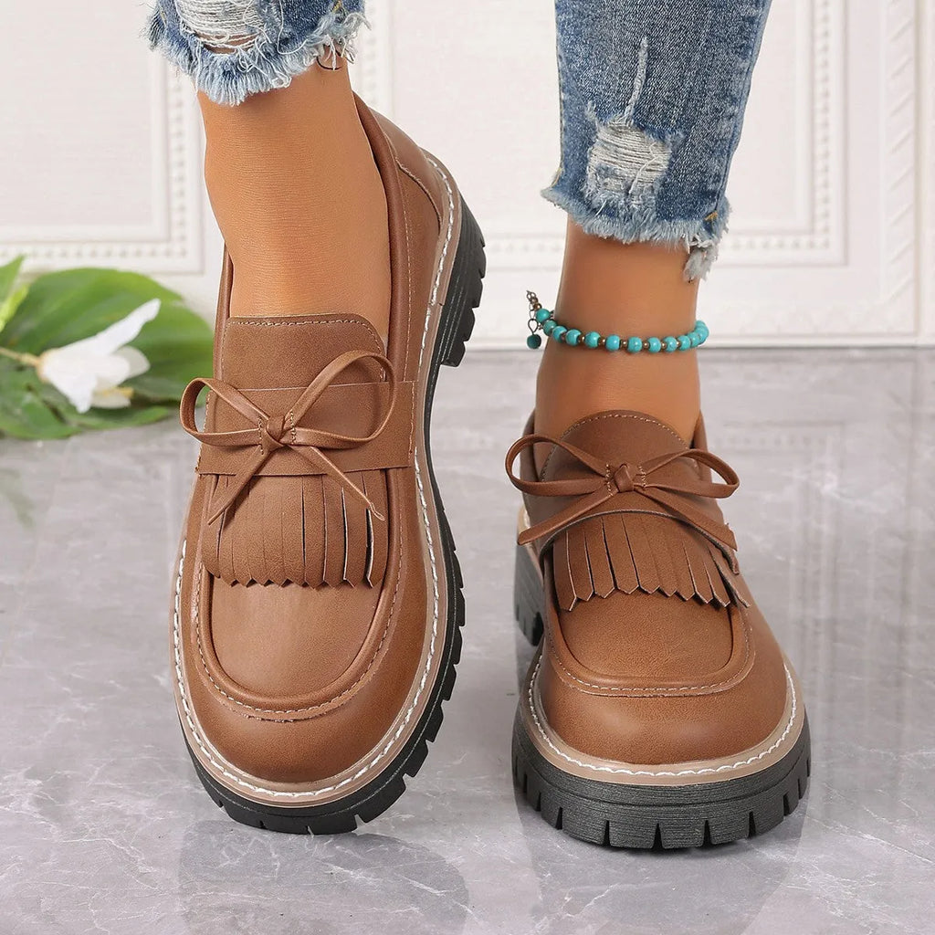 Women's black plus size loafers with tassel detail and a chunky rubber sole on a wooden floor.