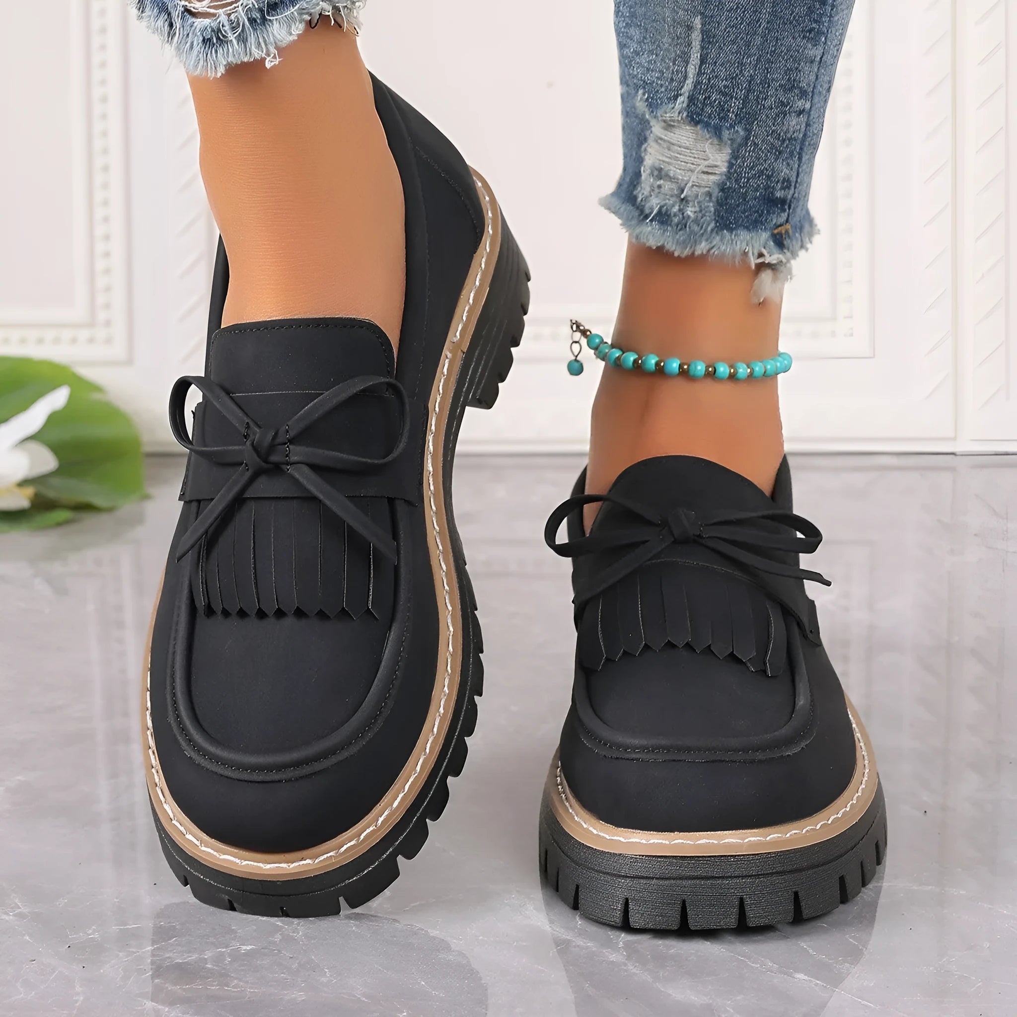 Women's black plus size loafers with tassel detail and a chunky rubber sole on a wooden floor.