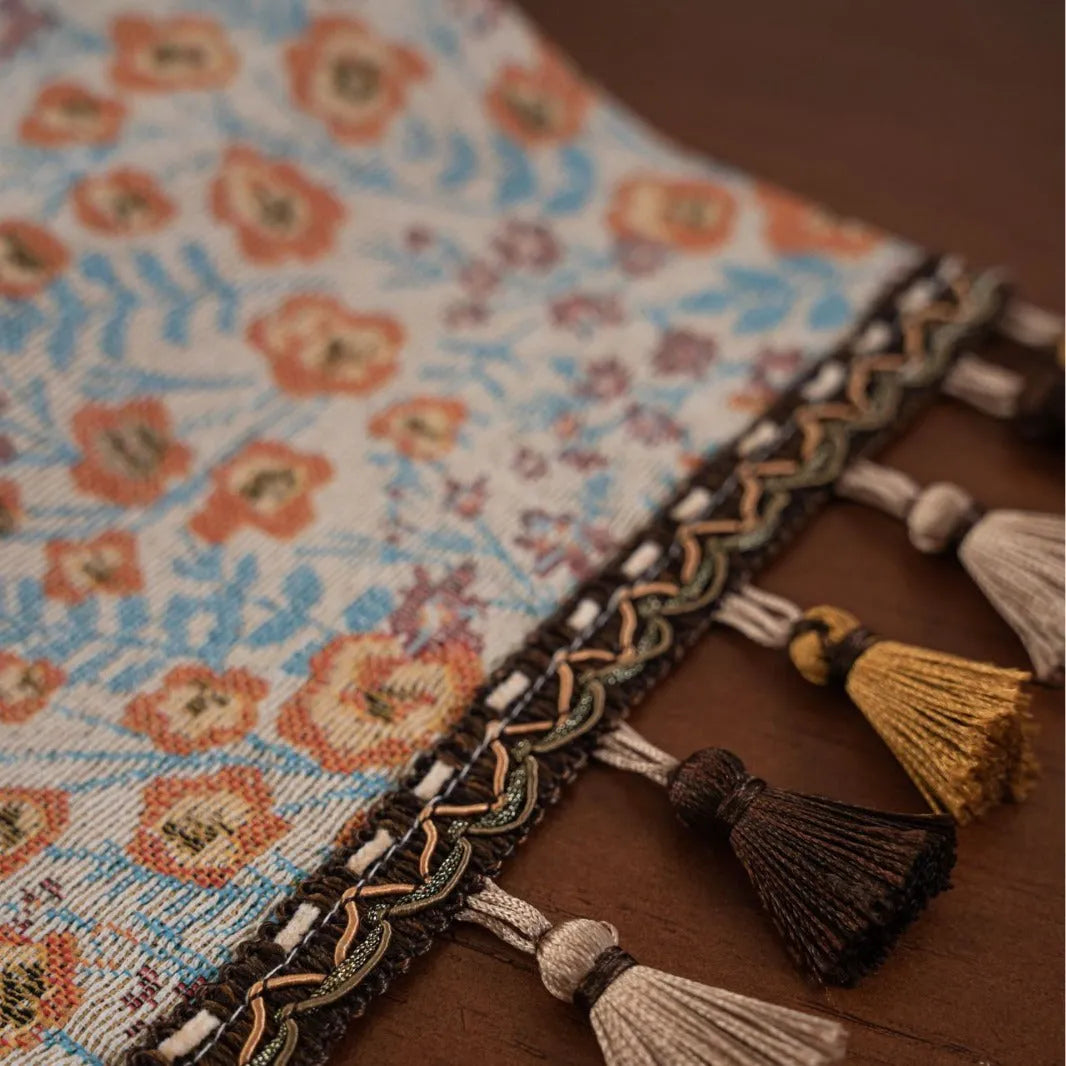 Beige jacquard table runner with floral pattern and long tassel fringes on a table.