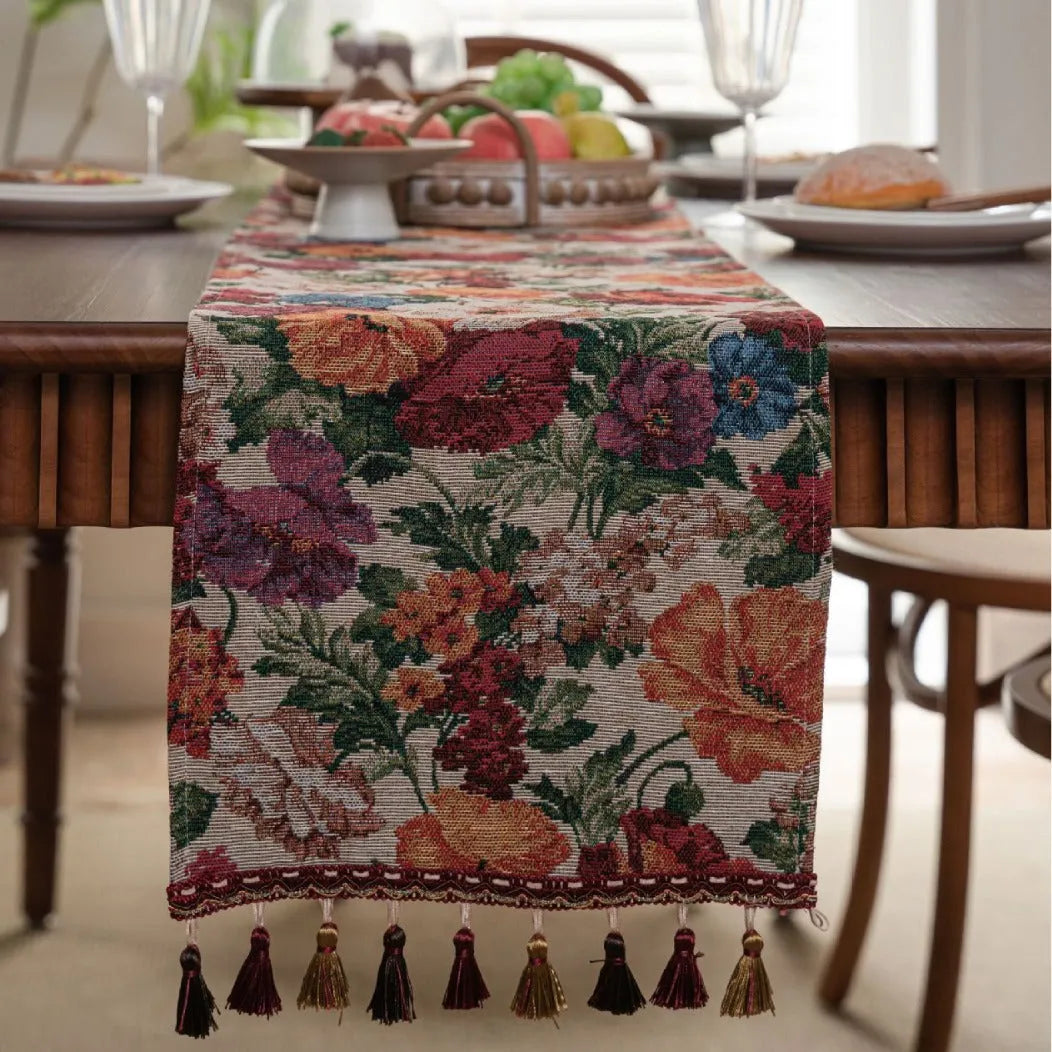 Beige jacquard table runner with floral pattern and long tassel fringes on a table.