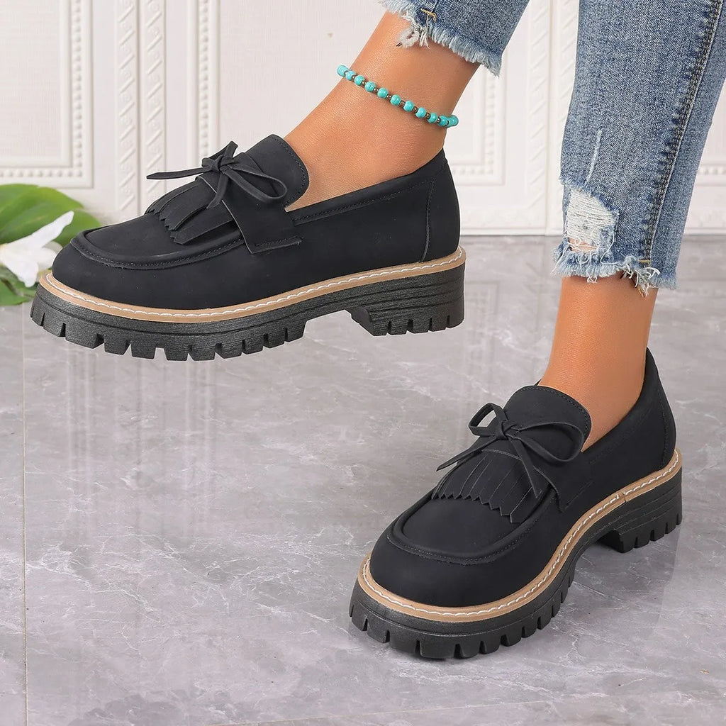 Women's black plus size loafers with tassel detail and a chunky rubber sole on a wooden floor.
