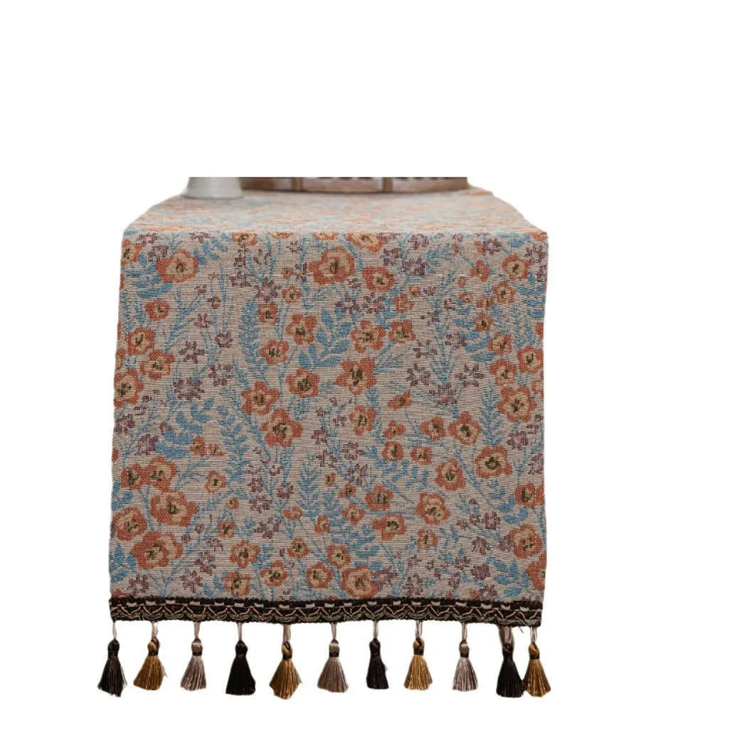Beige jacquard table runner with floral pattern and long tassel fringes on a table.