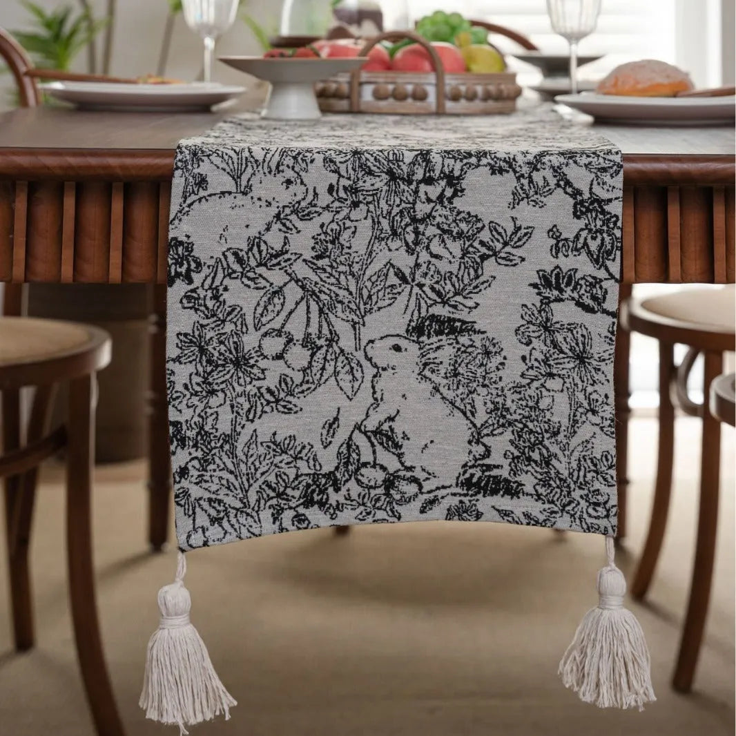 Beige jacquard table runner with floral pattern and long tassel fringes on a table.