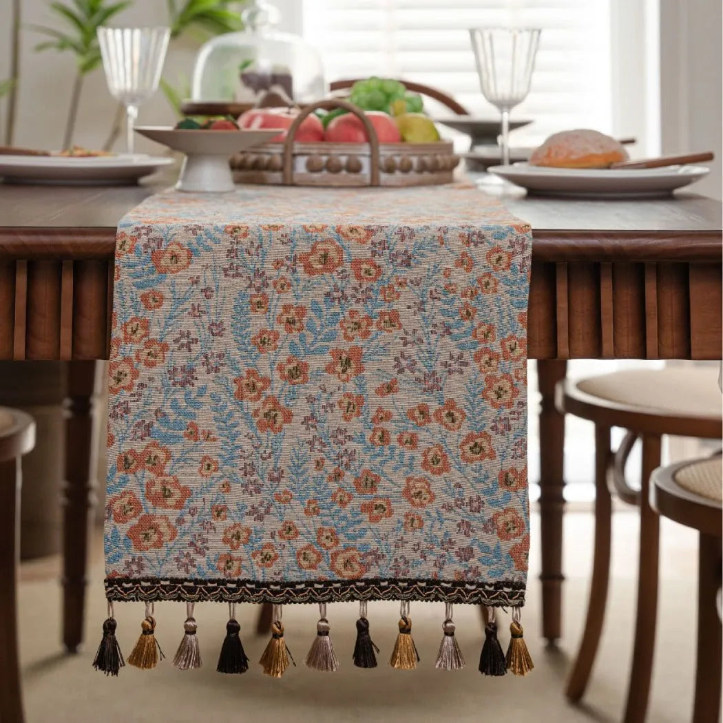 Beige jacquard table runner with floral pattern and long tassel fringes on a table.