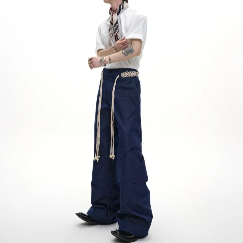 Men's dark blue straight-leg jeans with heavy 3D cutting, belt stitching, and tassel details on a loose fit.